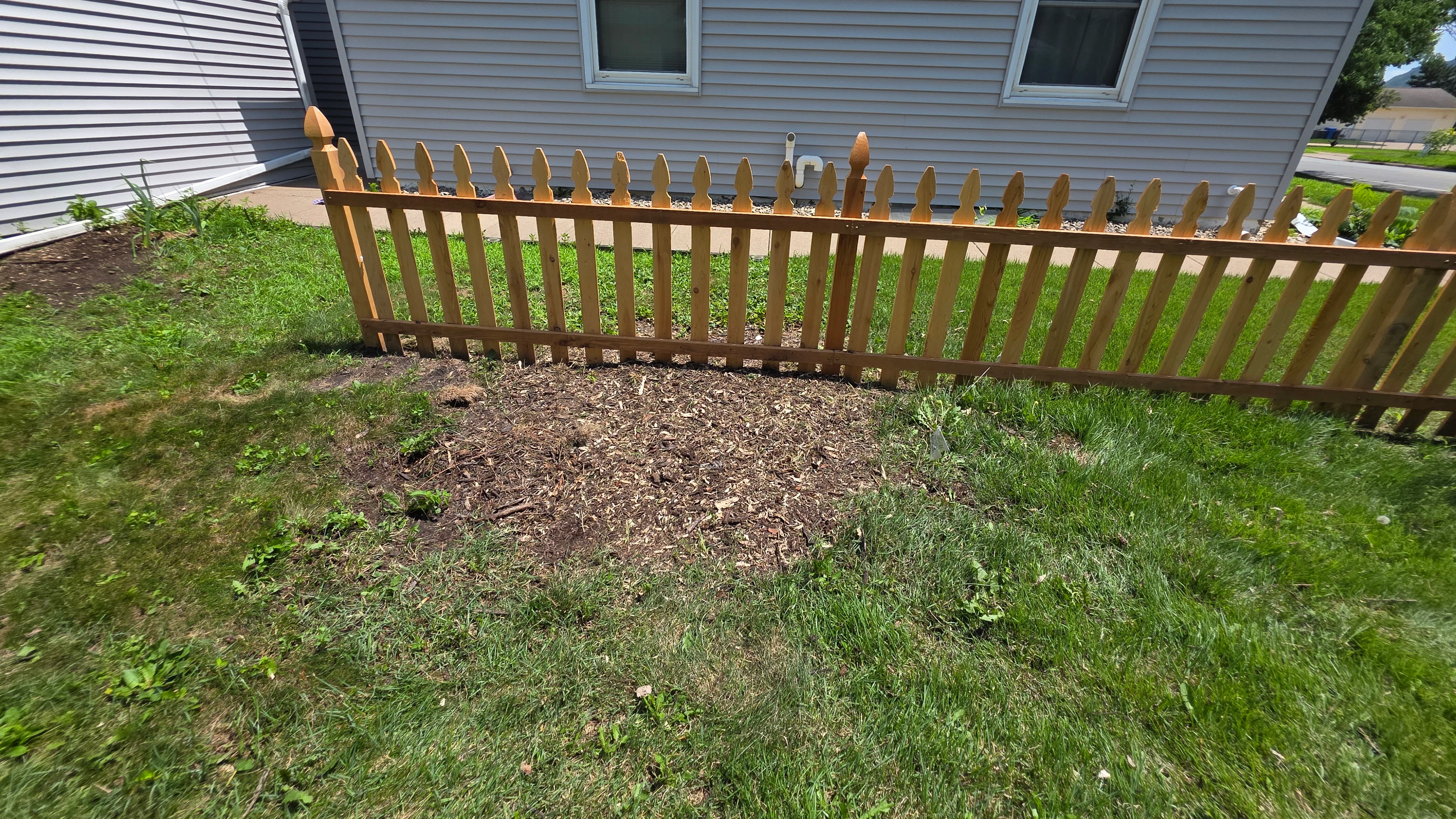 Natural wood picket fence with rounded tops installed along residential property with mulched landscape bed