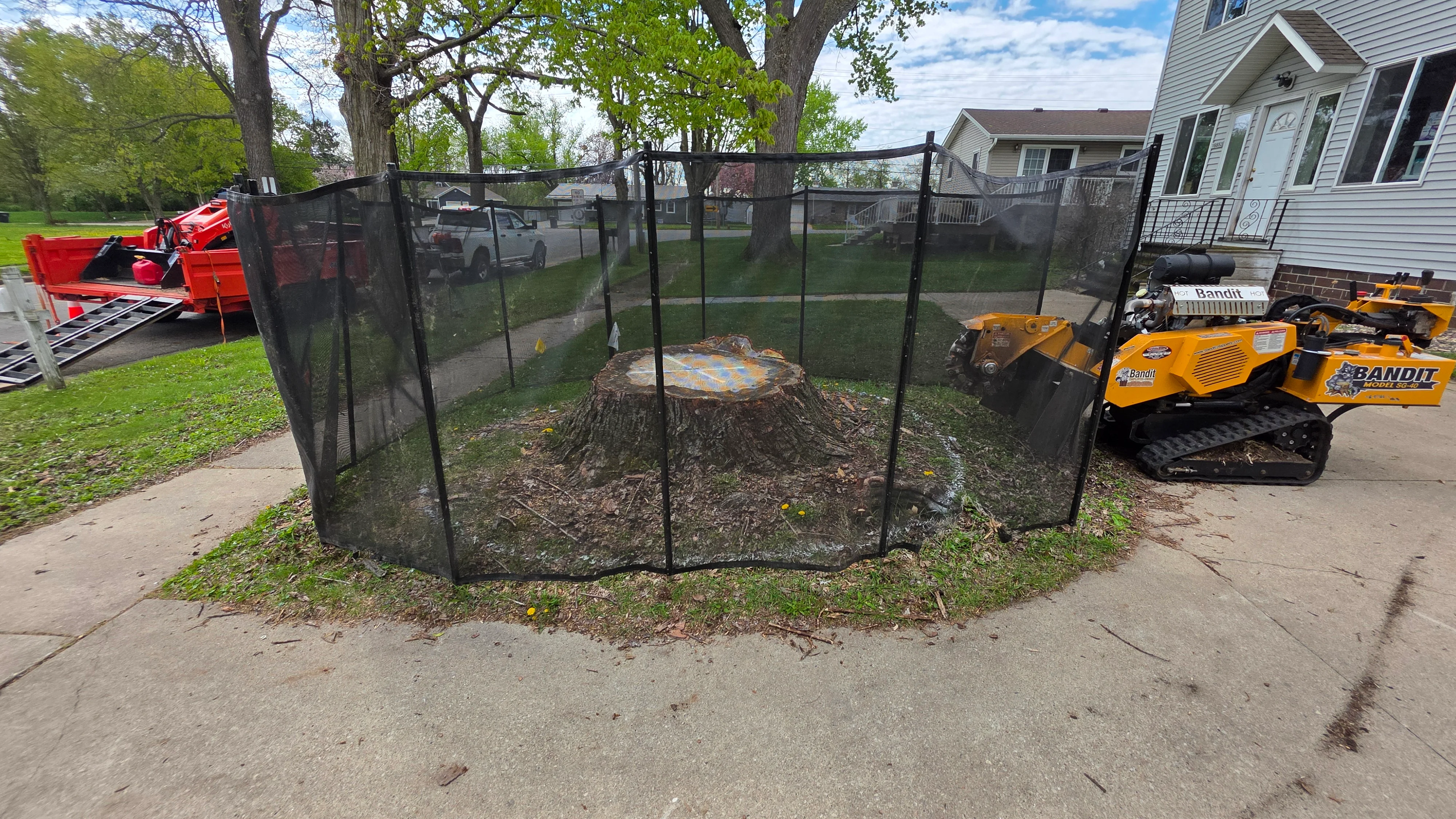 Large tree stump removal with black safety fence, cleared debris, and yellow Bandit equipment on residential driveway